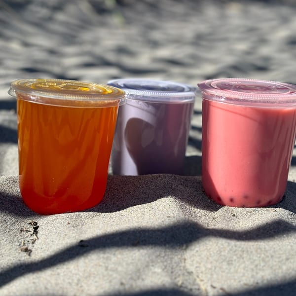Three colorful bubble tea drinks in plastic cups sit on a sandy beach under dappled sunlight.