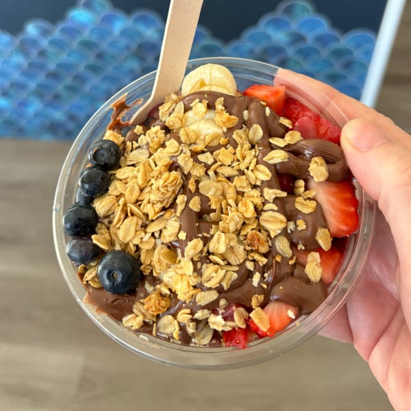 A hand holds a Gansett Poke acai bowl with chocolate, granola, blueberries, strawberries, and banana.